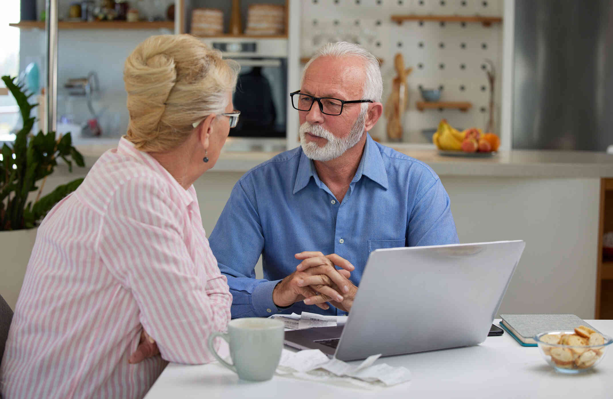 An elderly couple seated at a dining table, engaged in a serious conversation, surrounded by a laptop, notebook, and mugs.